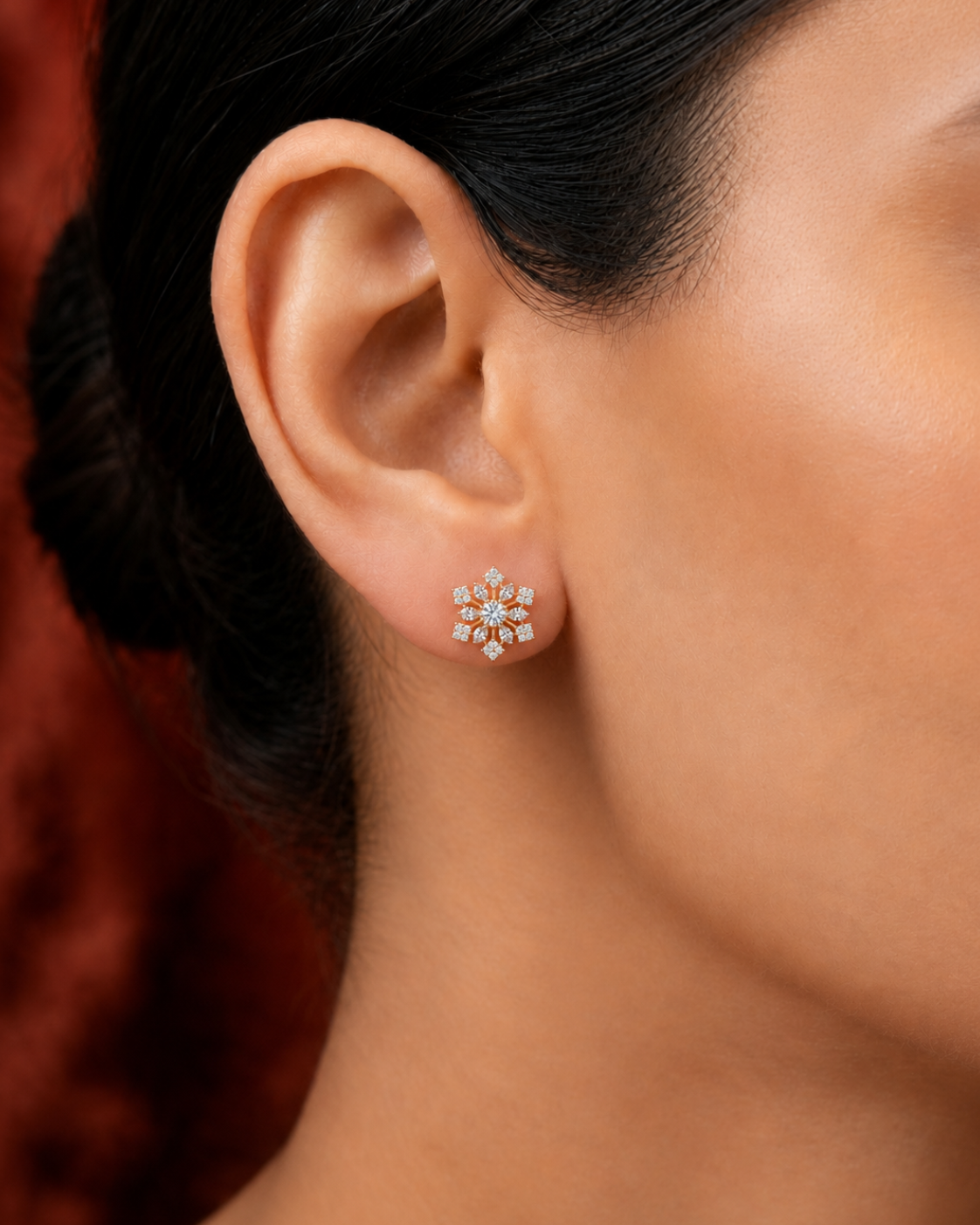 A close-up of a woman's ear wearing a small flower-shaped stud earring with clear gemstones in a warm metallic setting, against a dark red textured background.
