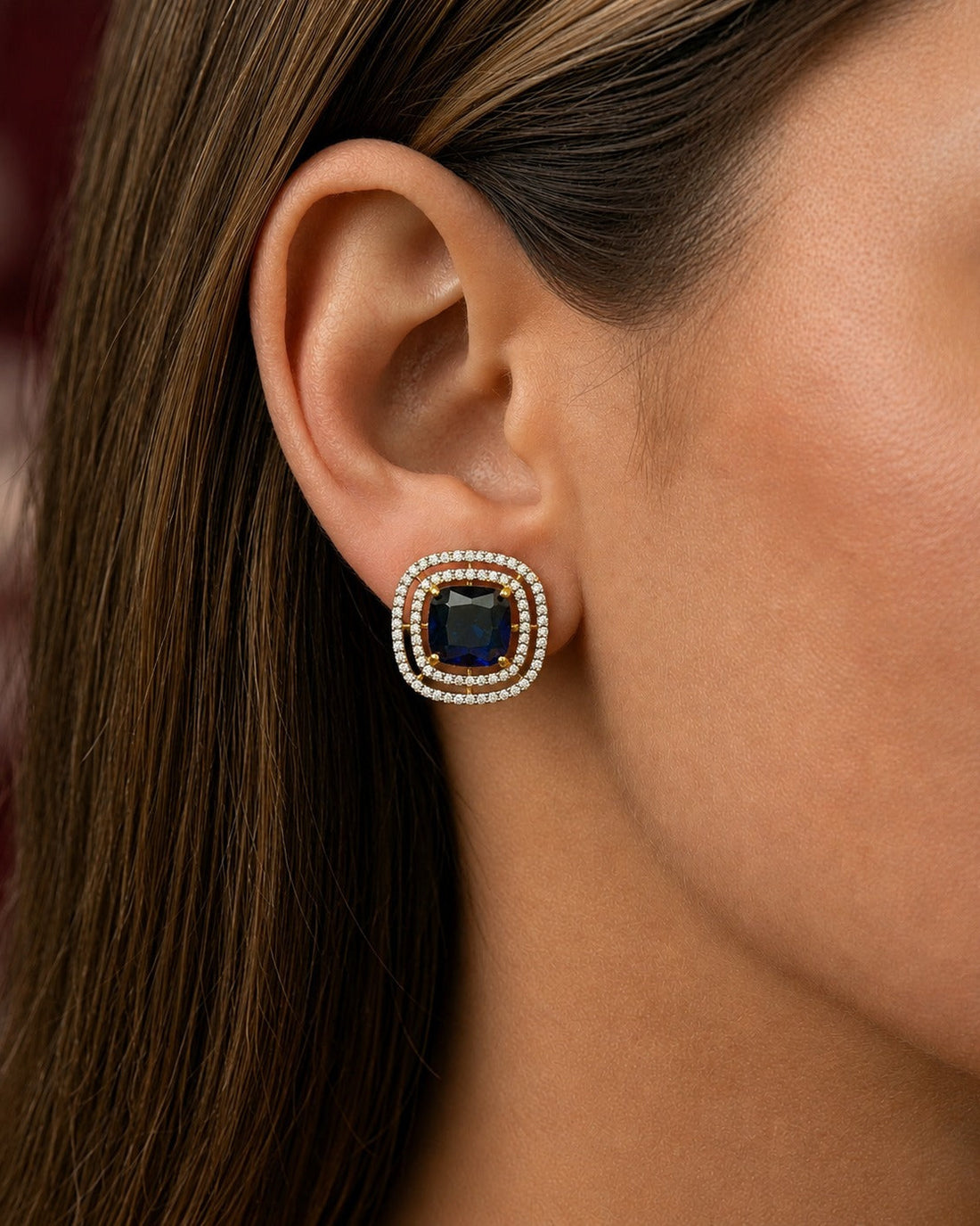 A close-up of a woman's ear wearing a square stud earring with a dark blue cushion-cut center stone surrounded by a double halo of small clear stones.