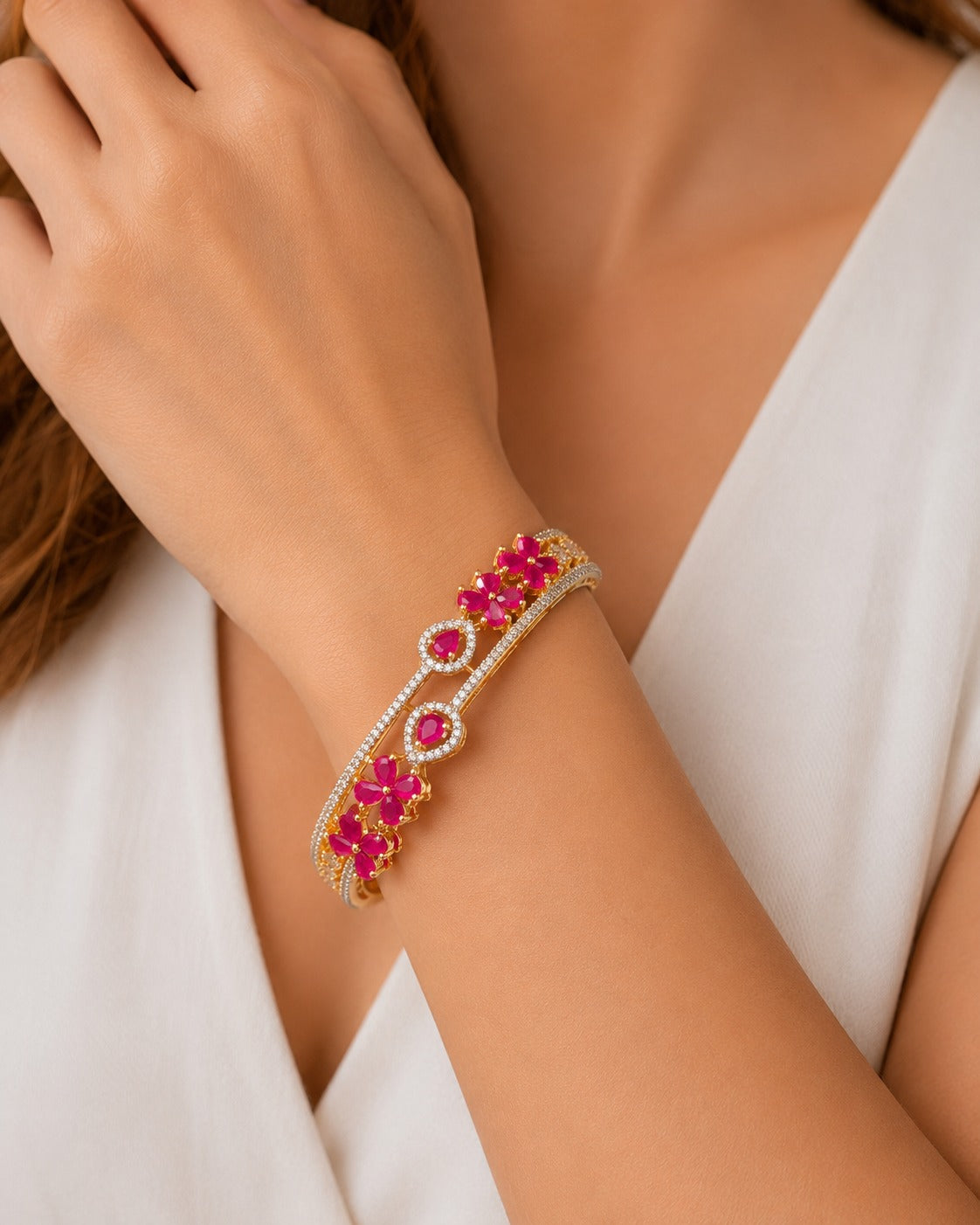 A close-up of a woman's wrist wearing a gold-plated bangle bracelet adorned with bright pink gemstones and small clear stones, paired with a white sleeveless top.
