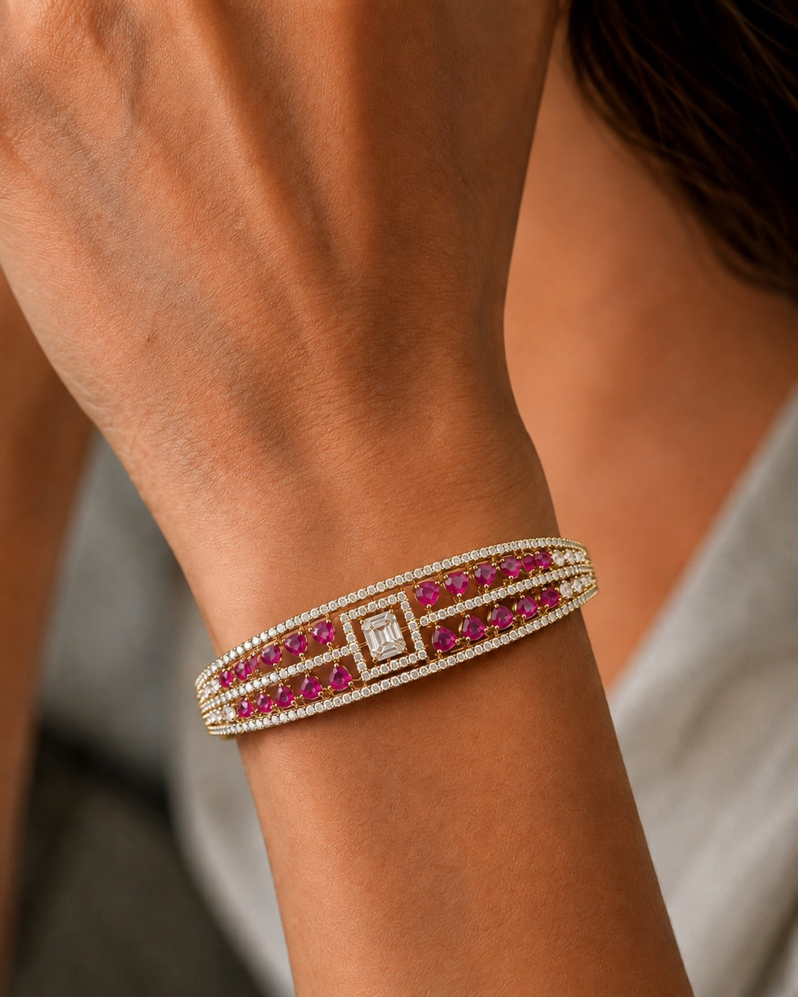 A close-up of a woman's wrist wearing a wide gold cuff bracelet adorned with rows of round pink gemstones and a central rectangular clear gemstone.