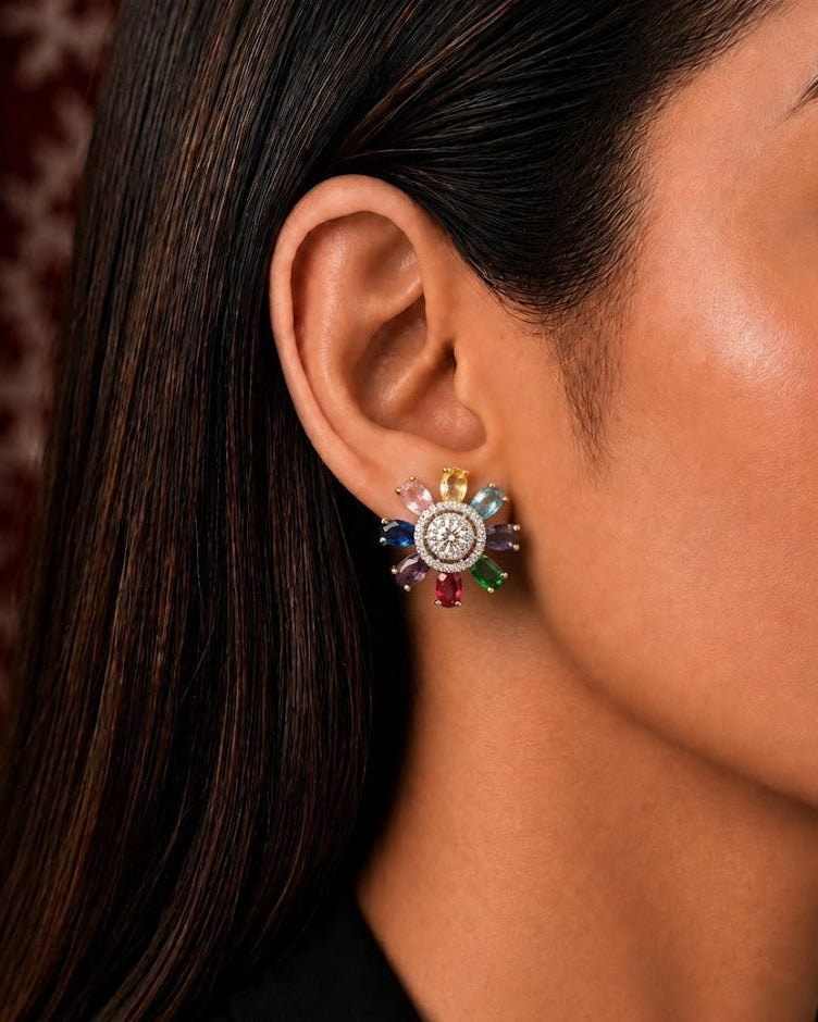A close-up side profile of a woman wearing a colorful, multi-stone flower-shaped stud earring with a silver-toned center and rainbow-colored petals.
