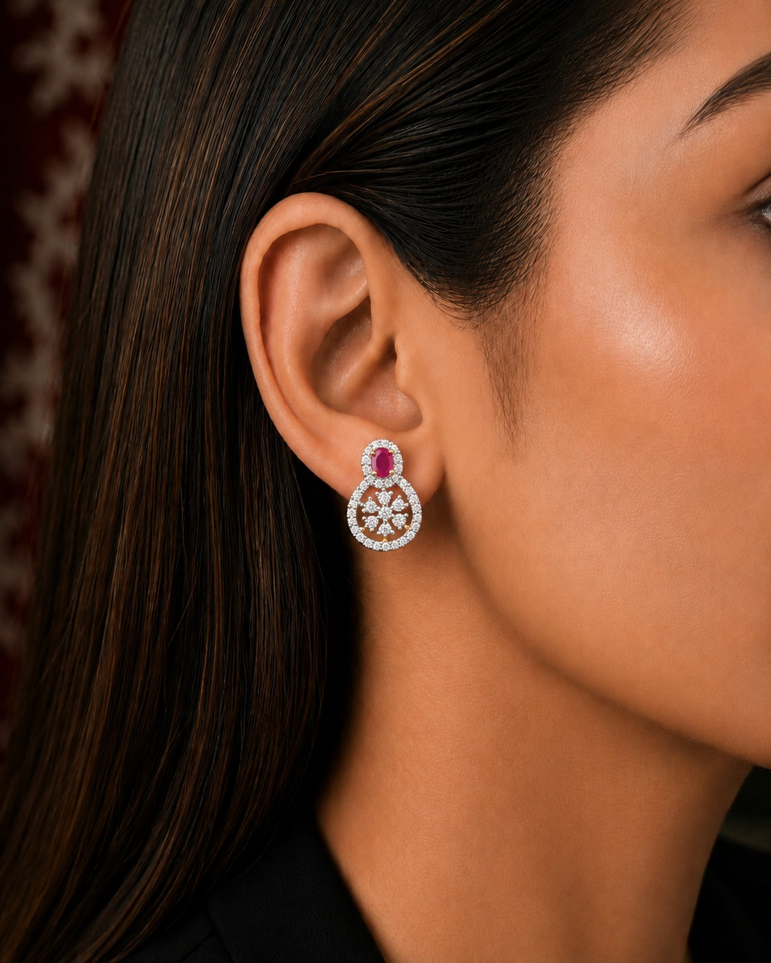 A close-up side profile of a woman wearing a single silver-toned dangle earring adorned with a central pink gemstone and a surrounding halo of small clear stones.