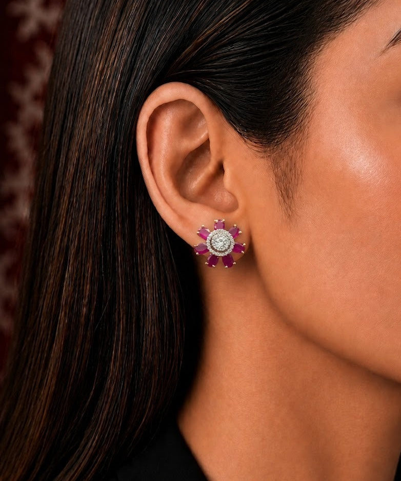 A close-up side profile of a woman wearing a single floral stud earring. The earring features a central clear round stone surrounded by a halo of small pinkish-red stones and a silver-toned setting.