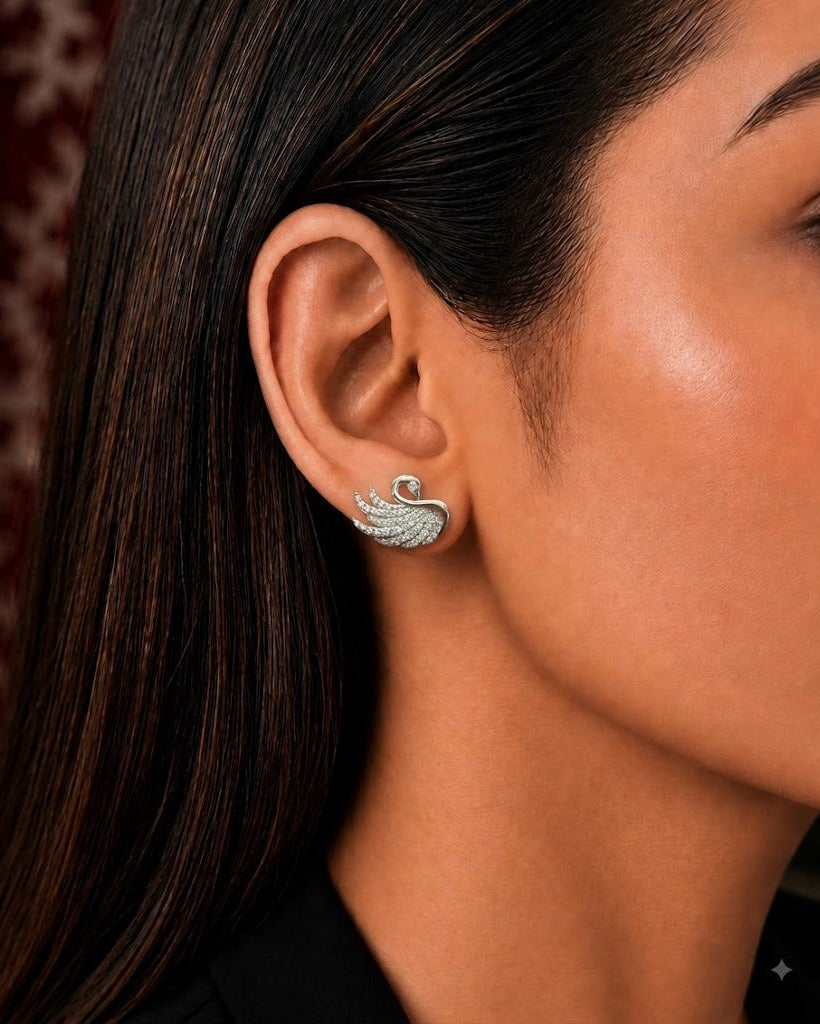 A close-up side profile of a woman wearing a small silver-toned swan-shaped stud earring with a clear gemstone, shown against her ear and dark hair.