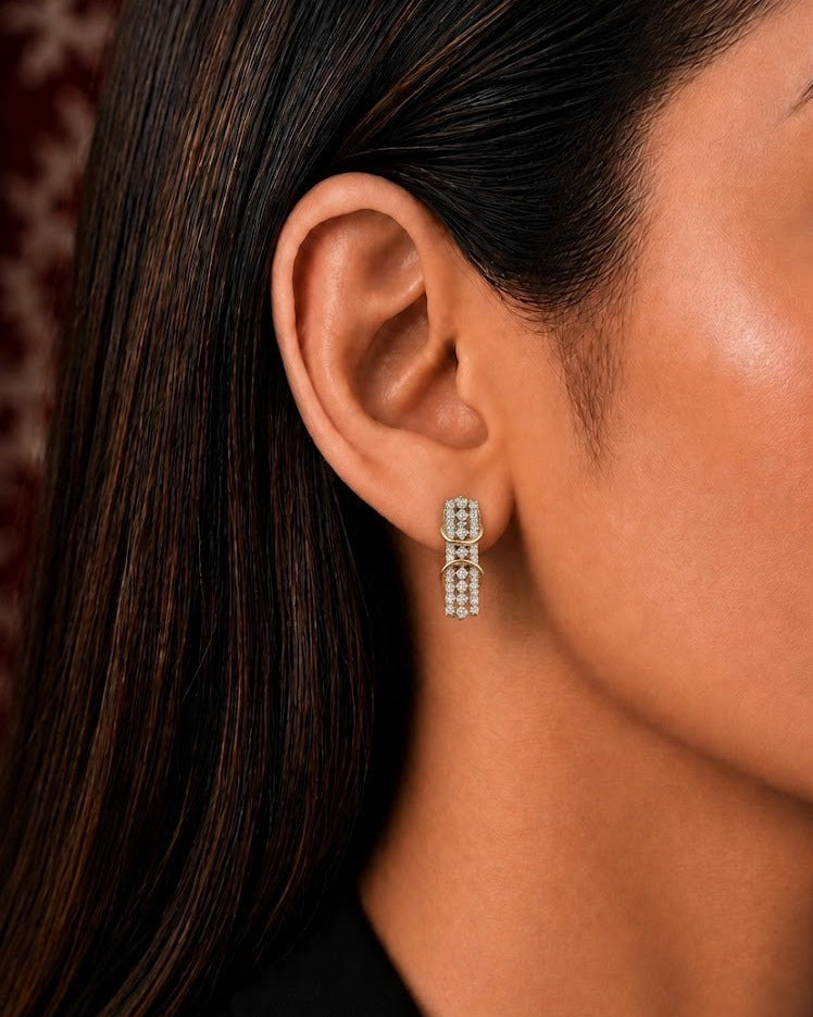 A close-up side profile of a woman wearing a small vertical drop earring with multiple clear stones set in a gold-tone frame.