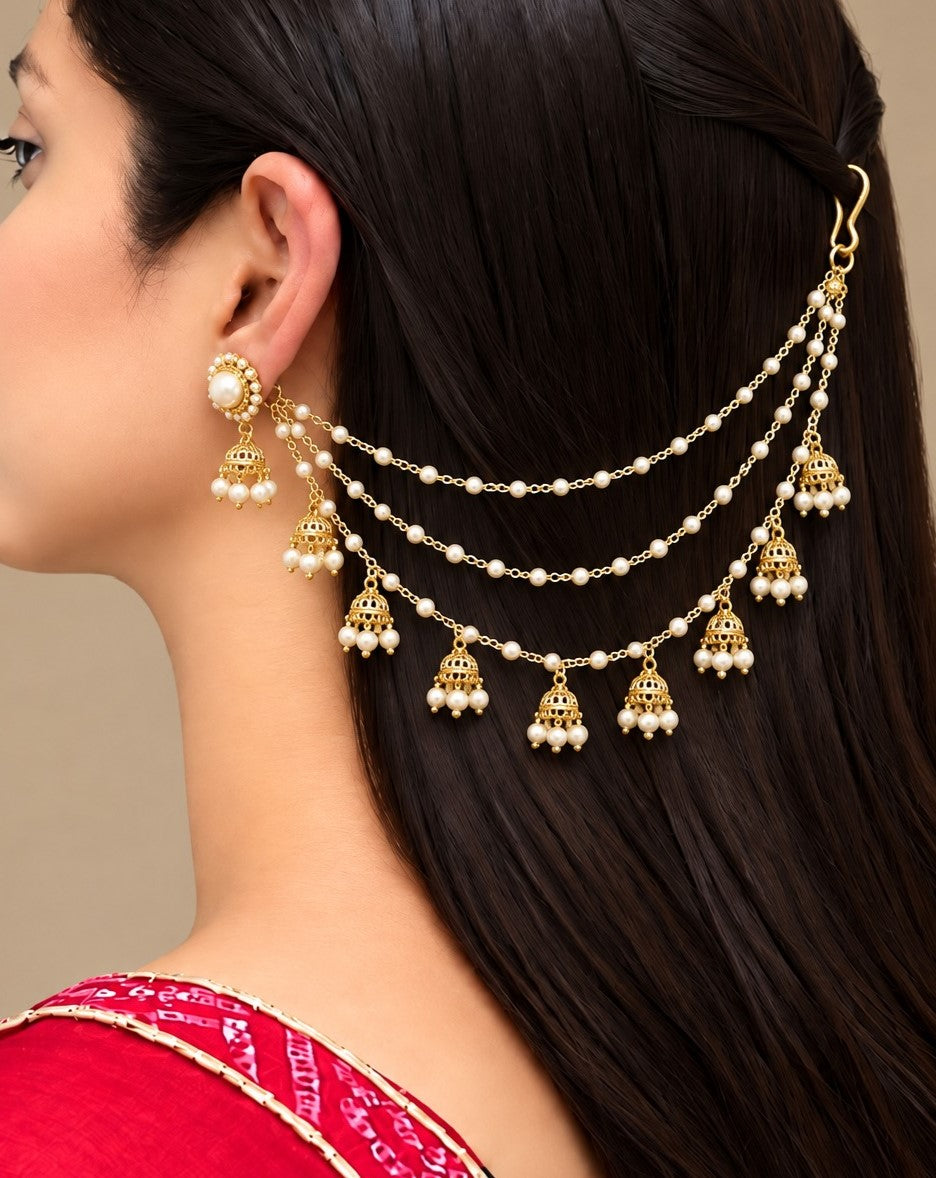 A close-up side profile of a woman with long dark hair wearing a gold and pearl hair chain accessory with matching dangling earrings. The jewelry features multiple strands of small white pearls and ornate gold bell-shaped charms.