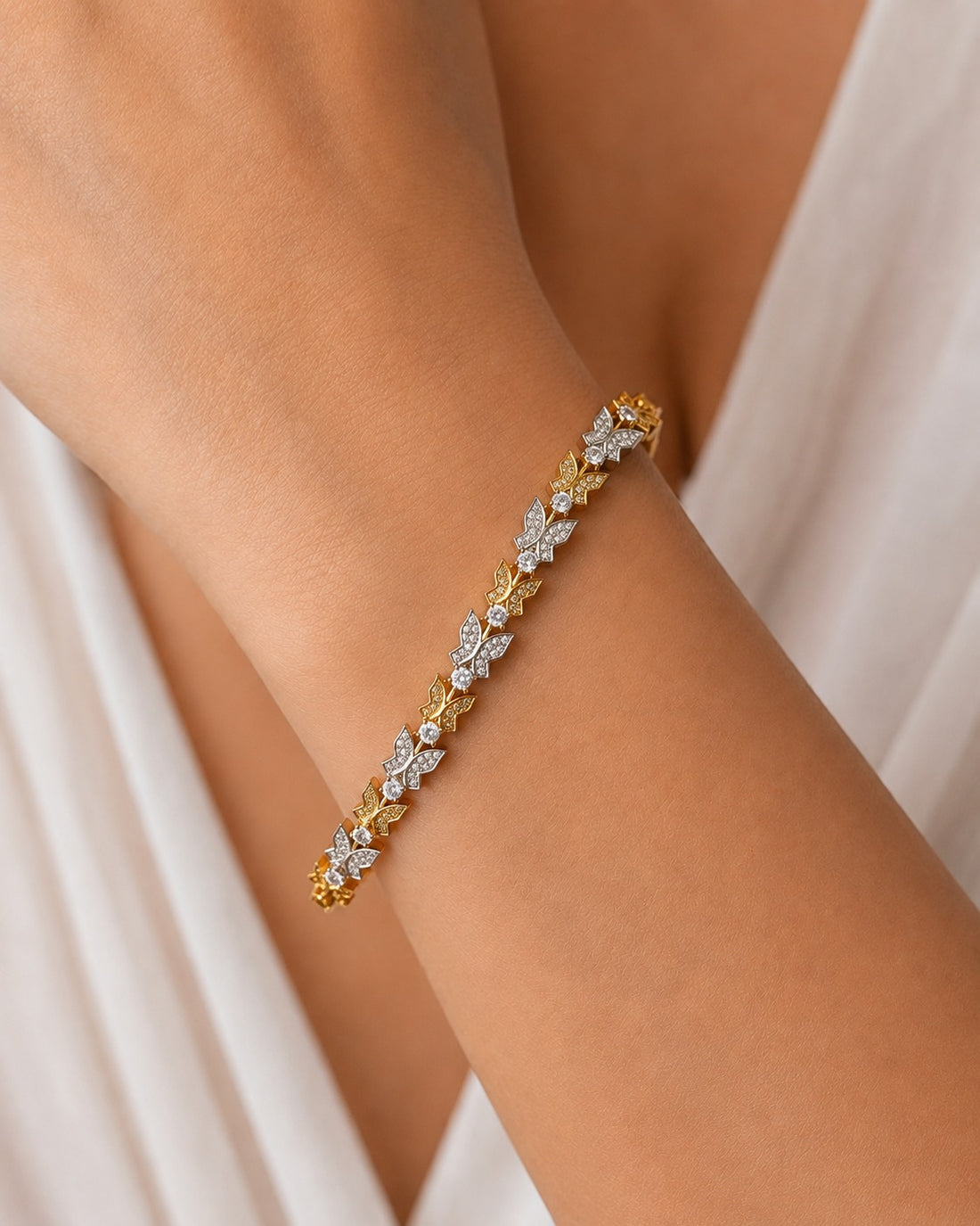 A delicate gold bracelet with diamond-encrusted floral motifs worn on a person's wrist, set against a soft white draped garment.