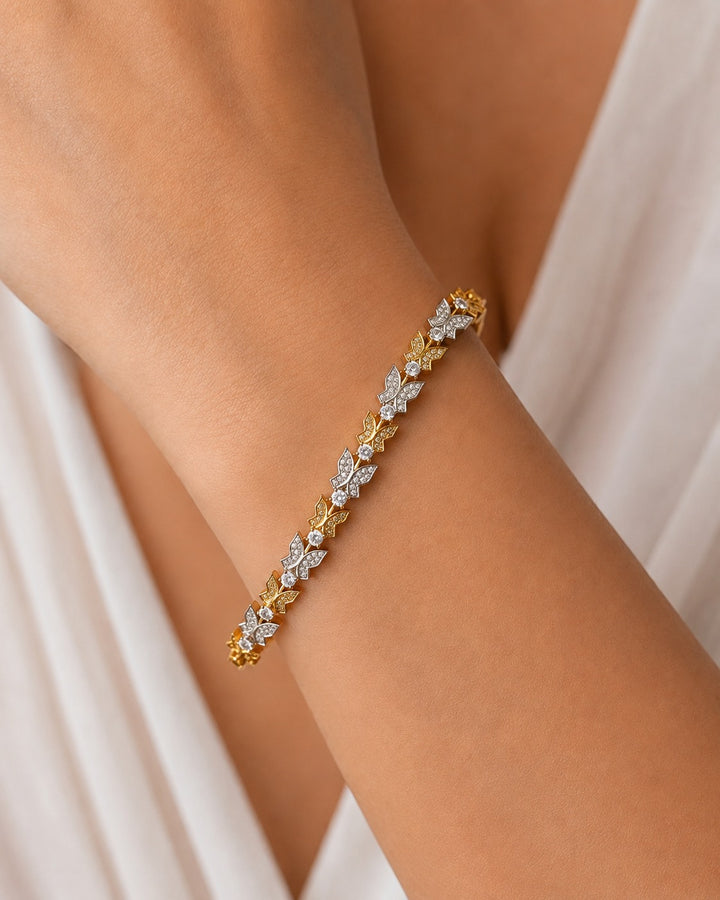 A delicate gold bracelet with diamond-encrusted floral motifs worn on a person's wrist, set against a soft white draped garment.