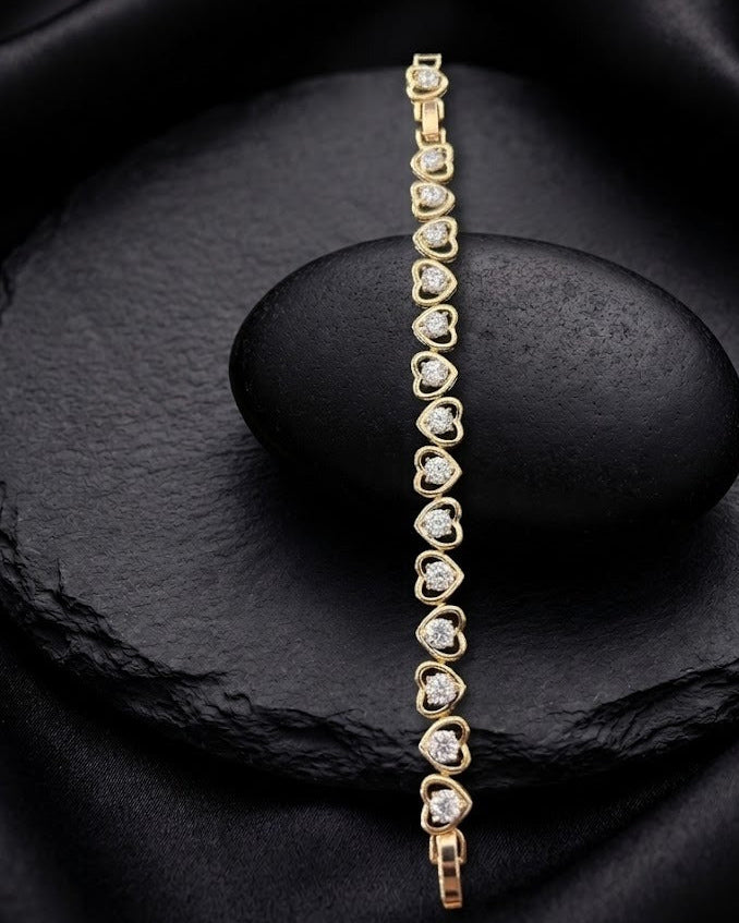 A delicate gold bracelet with small heart-shaped clear stones, displayed vertically on a dark stone pedestal against a black satin fabric background.