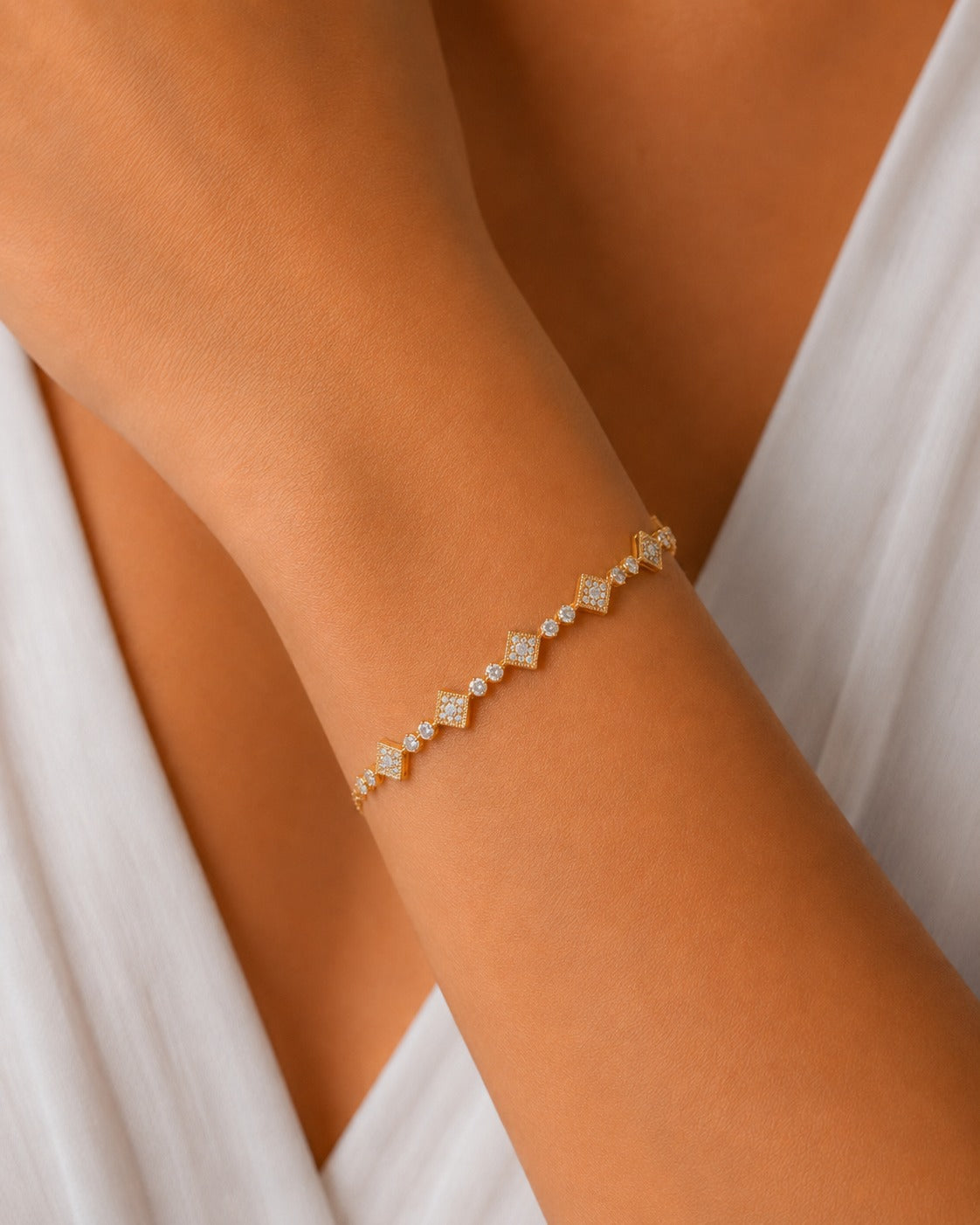 A delicate gold chain bracelet with small clear stones set in diamond-shaped links, worn on a person's wrist against a white draped garment.