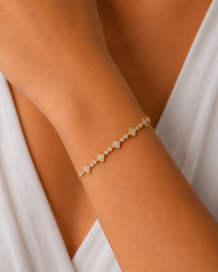 A delicate gold chain bracelet with small clear stones set in diamond-shaped links, worn on a person's wrist against a white draped garment.