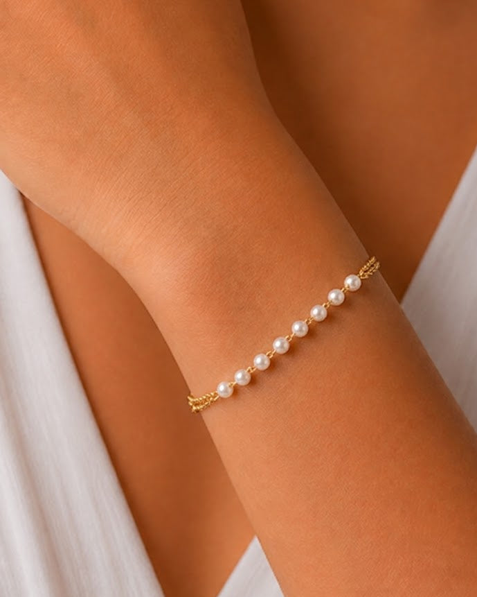 A delicate gold chain bracelet with a row of small white pearls, shown worn on a person's wrist against a white dress.