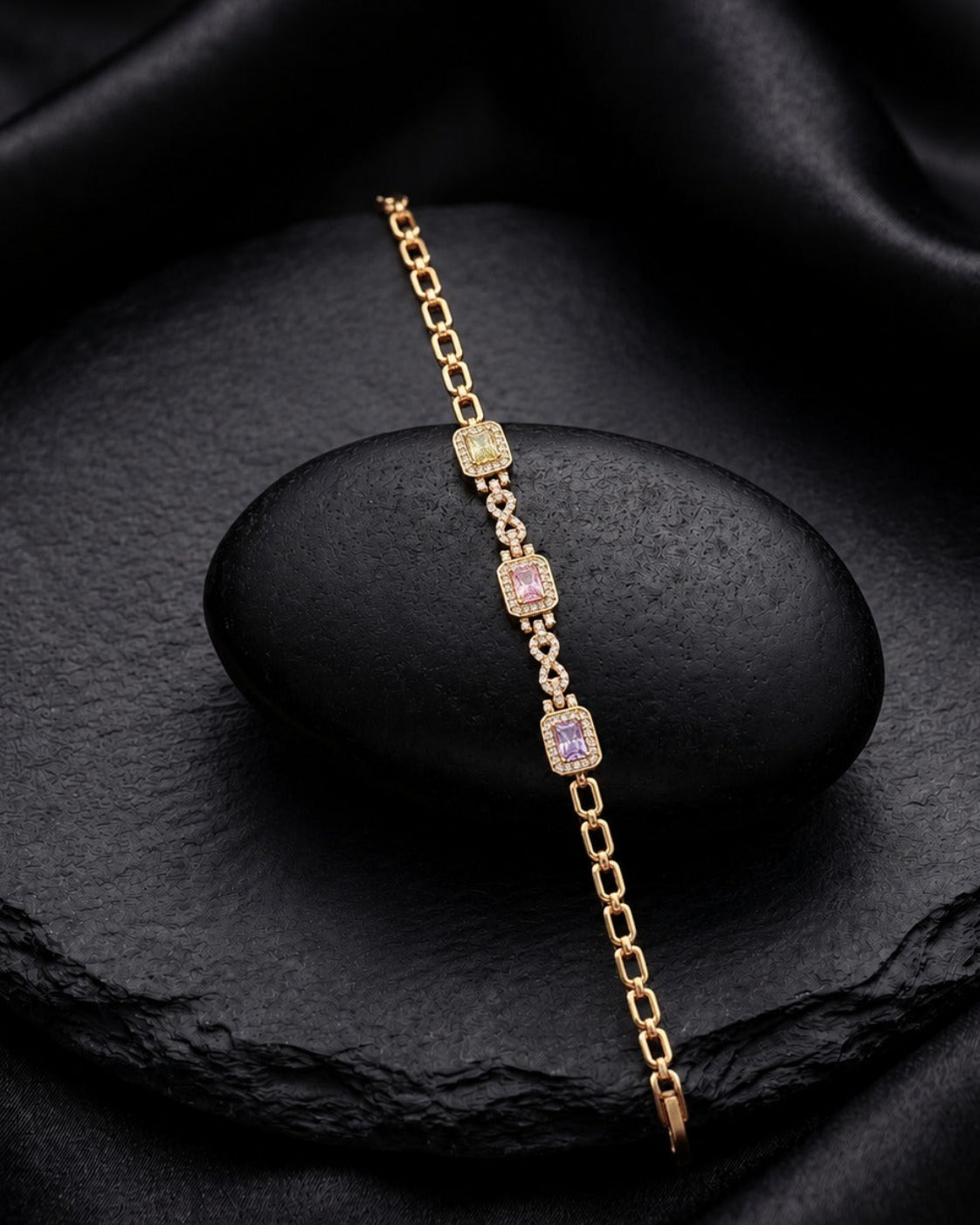 A delicate gold chain bracelet with three rectangular purple gemstone accents, displayed on a black stone pedestal against a draped black satin background.