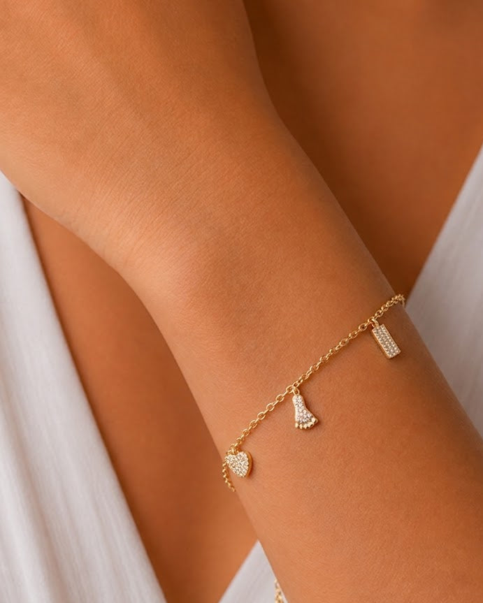 A delicate gold chain bracelet with three small charms (a heart, a fan, and a rectangular pavé piece) worn on a model's wrist.