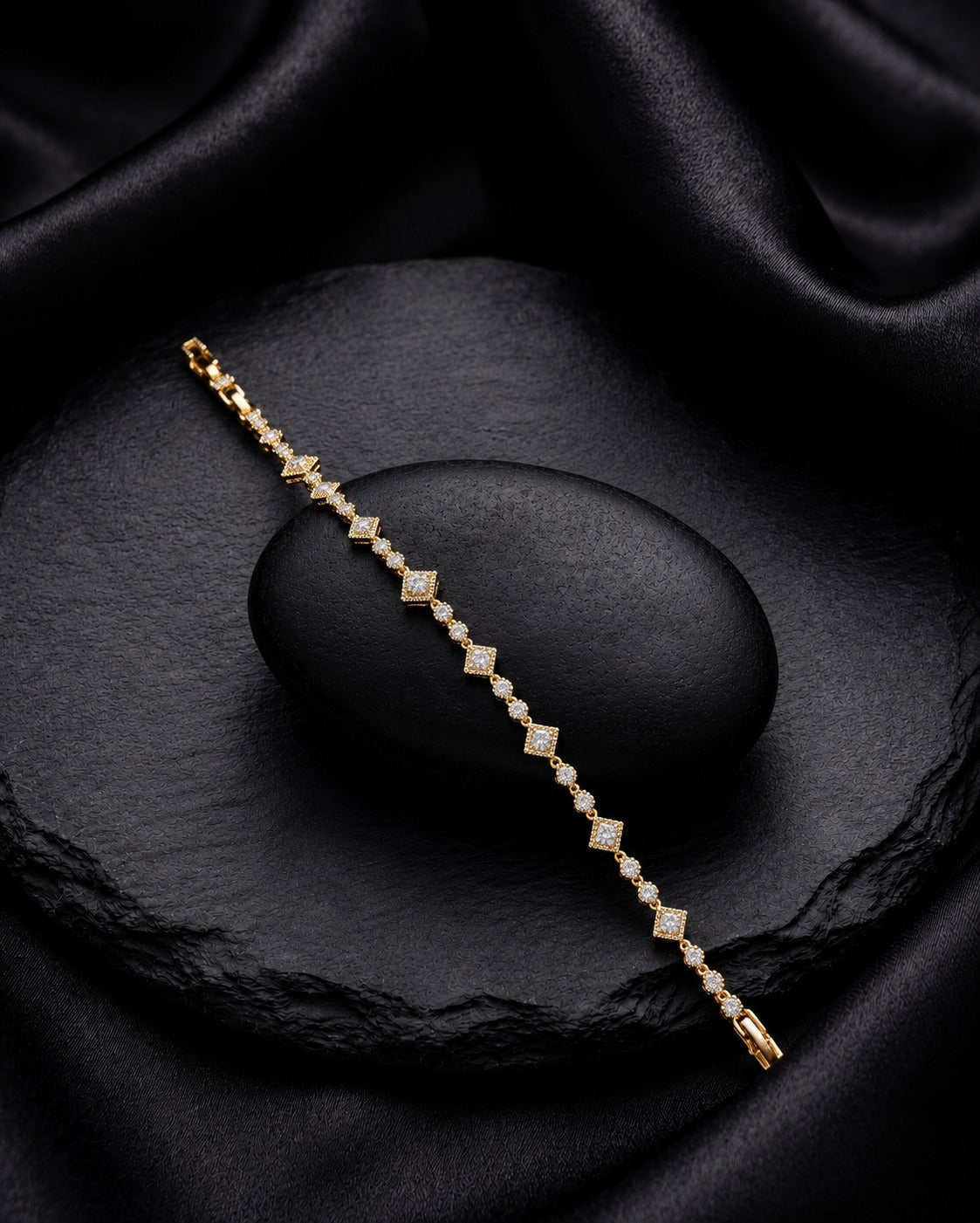 A delicate gold tennis bracelet with small clear stones set in a repeating diamond pattern, displayed diagonally on a dark textured stone over black satin fabric.