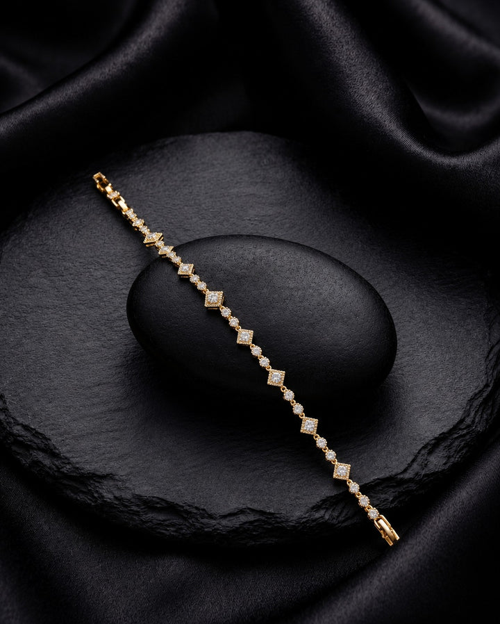 A delicate gold tennis bracelet with small clear stones set in a repeating diamond pattern, displayed diagonally on a dark textured stone over black satin fabric.
