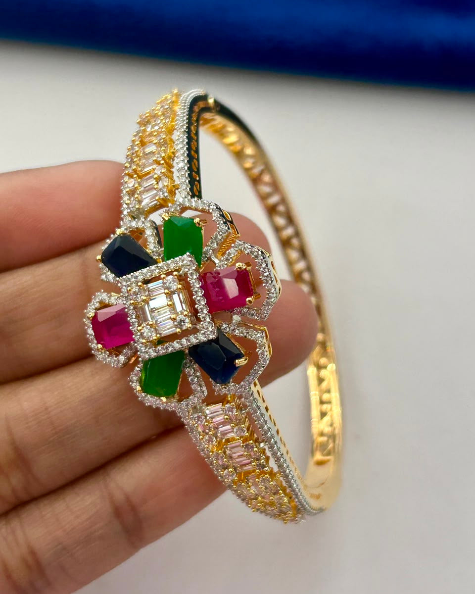 A gold-plated bangle bracelet with a large floral centerpiece, adorned with colorful gemstones including pink, green, and blue, held in a person's hand against a white background with a blue velvet surface in the background.