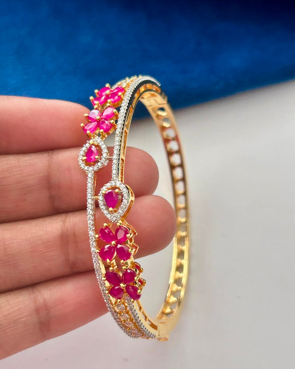 A gold-plated bangle bracelet decorated with bright pink gemstones and pavé-set clear stones, held in a person's hand against a blue and white background.