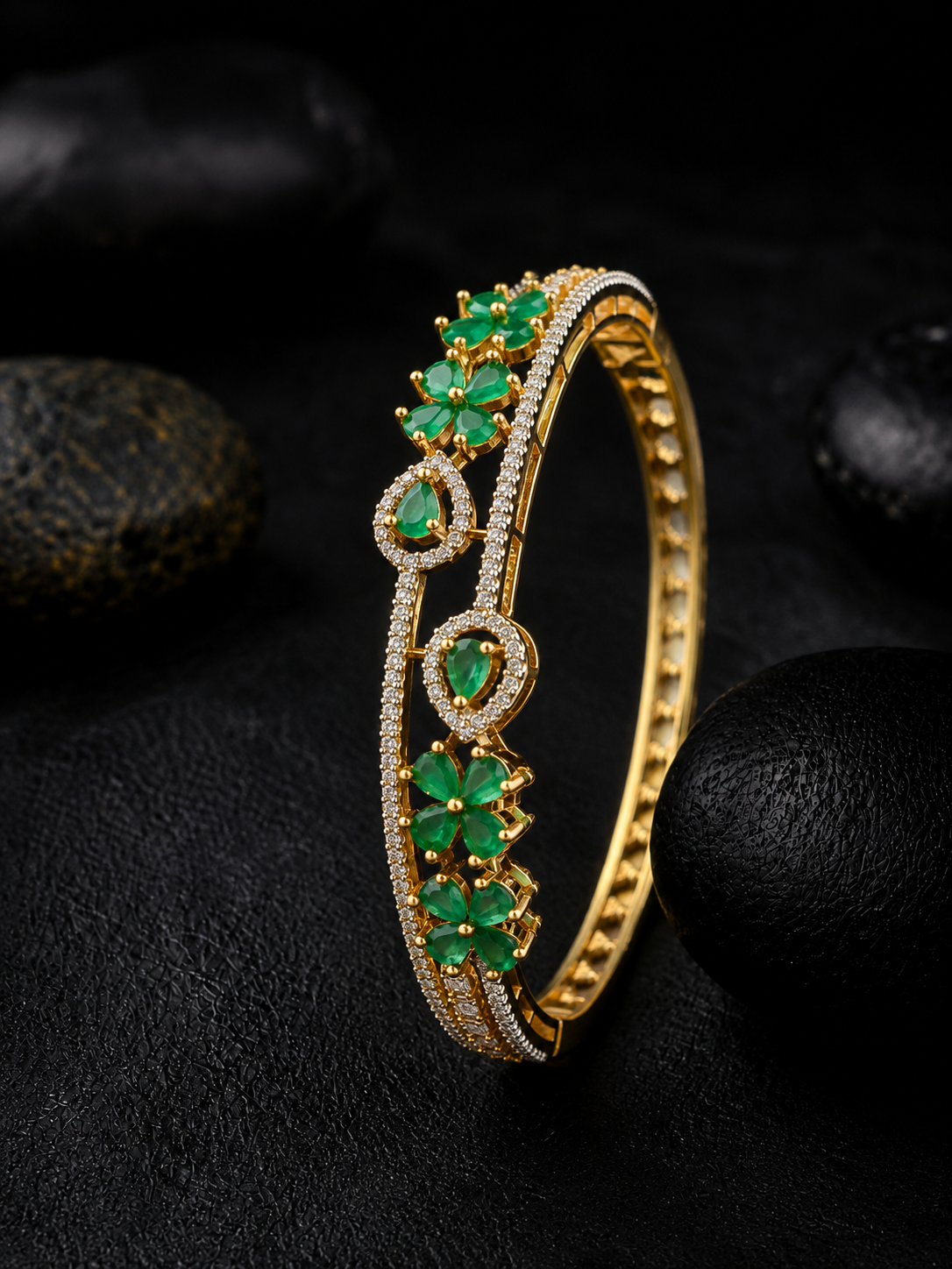 A gold-plated bangle bracelet decorated with green emerald-cut gemstones and pavé-set clear stones, displayed on a dark textured surface with black stones in the background.