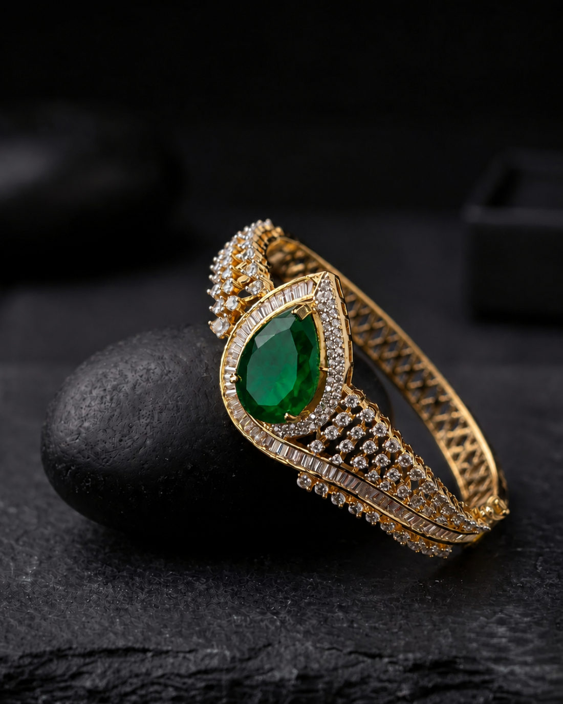 A gold-plated bangle bracelet featuring a large pear-cut green emerald center stone and rows of small clear stones, displayed on a dark textured surface.
