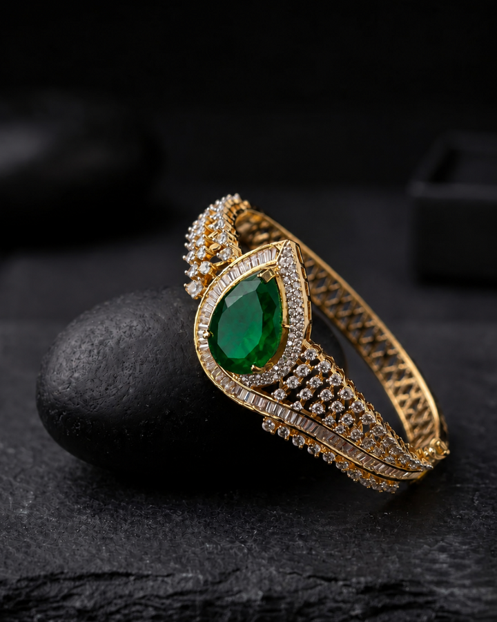 A gold-plated bangle bracelet featuring a large pear-cut green emerald center stone and rows of small clear stones, displayed on a dark textured surface.