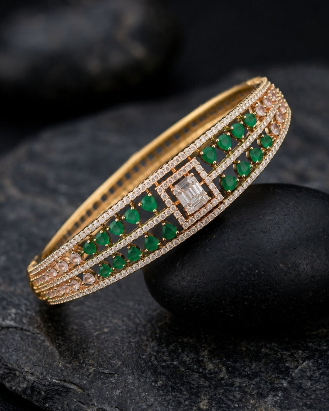 A gold-plated bangle bracelet featuring rows of green emerald-cut gemstones and small clear stones, displayed on a dark stone surface.