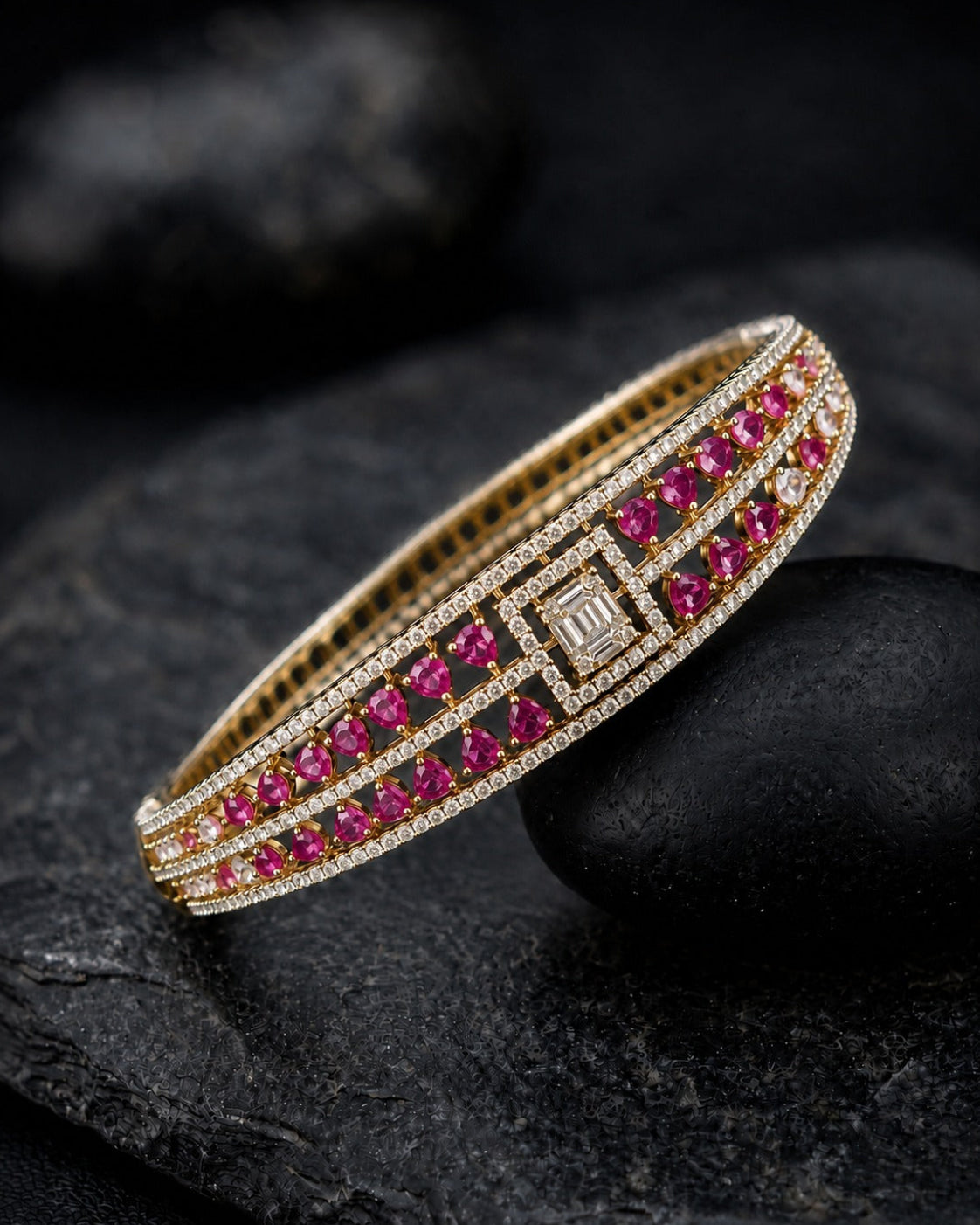 A gold-plated bangle bracelet featuring rows of vibrant pinkish-red round-cut stones and a central square-cut stone, accented with small clear pavé stones, displayed on a dark textured stone surface.