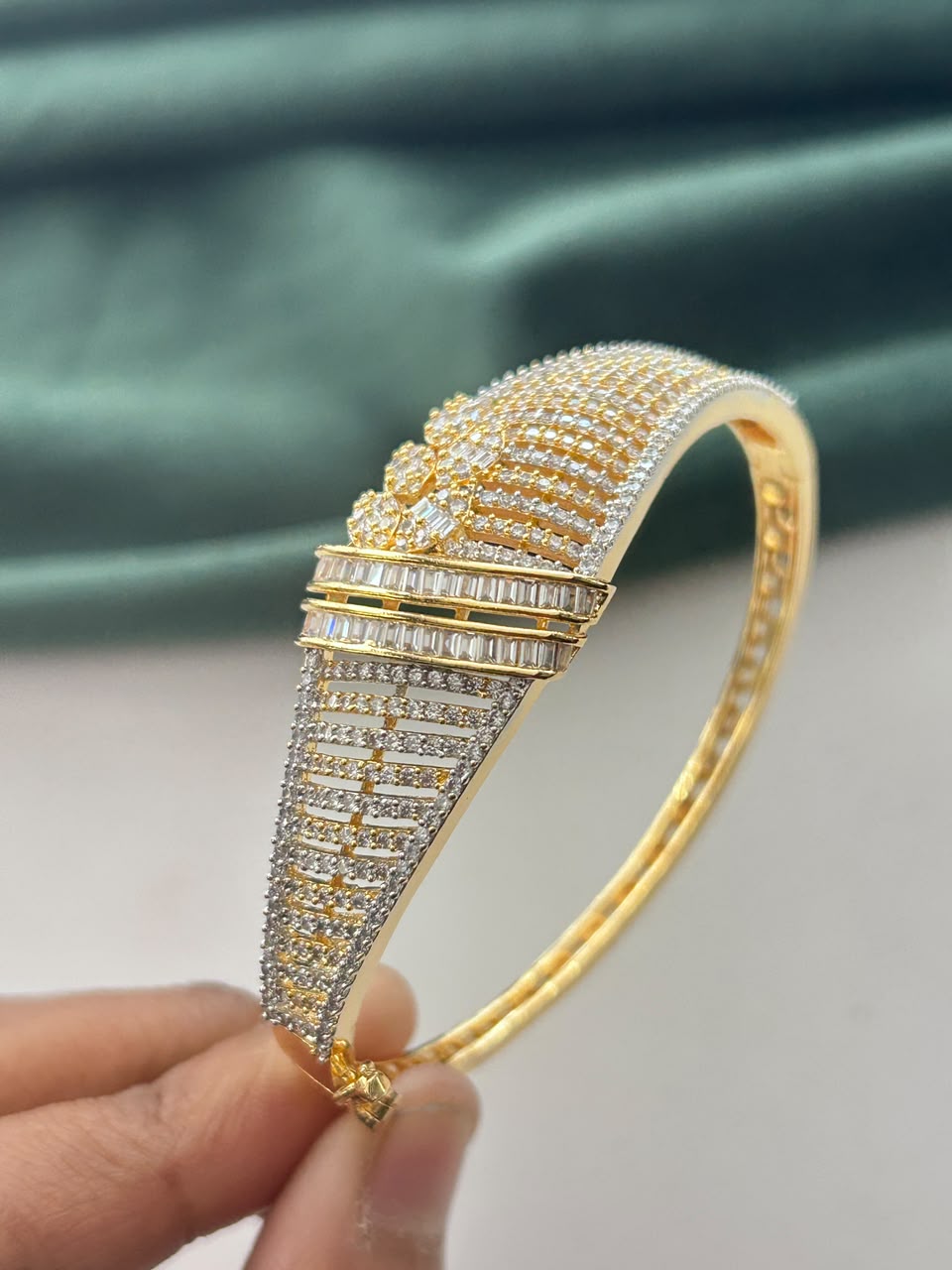 A gold-plated bangle bracelet heavily embellished with pave-set clear stones, held by a hand against a soft teal fabric background.