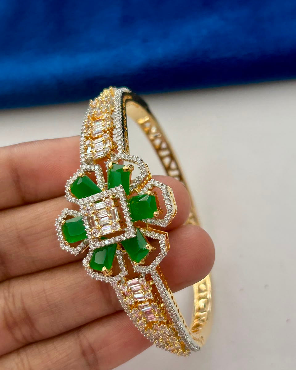 A gold-plated bangle bracelet with a central floral motif featuring green emerald-cut stones and clear baguette and round stones, held in a person's hand against a blue and white background.