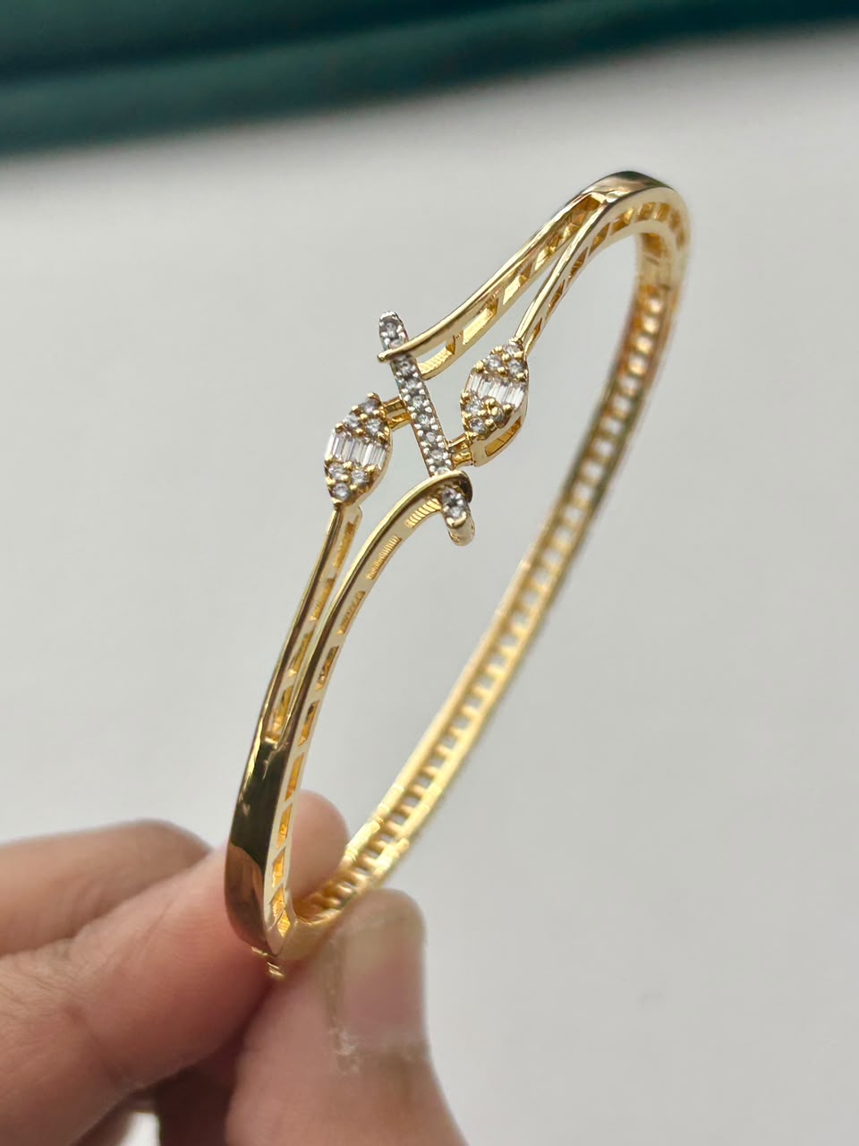 A gold-plated bangle bracelet with a decorative cross and small clear stones, held by a hand against a light gray background.