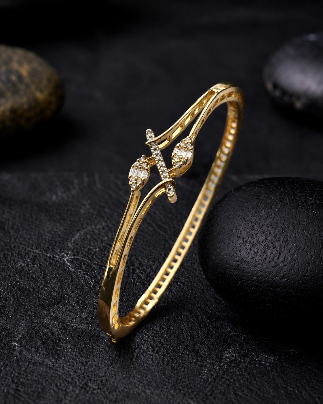 A gold-plated bangle bracelet with a decorative cross motif and small clear stones, displayed on a dark textured surface with black stones.