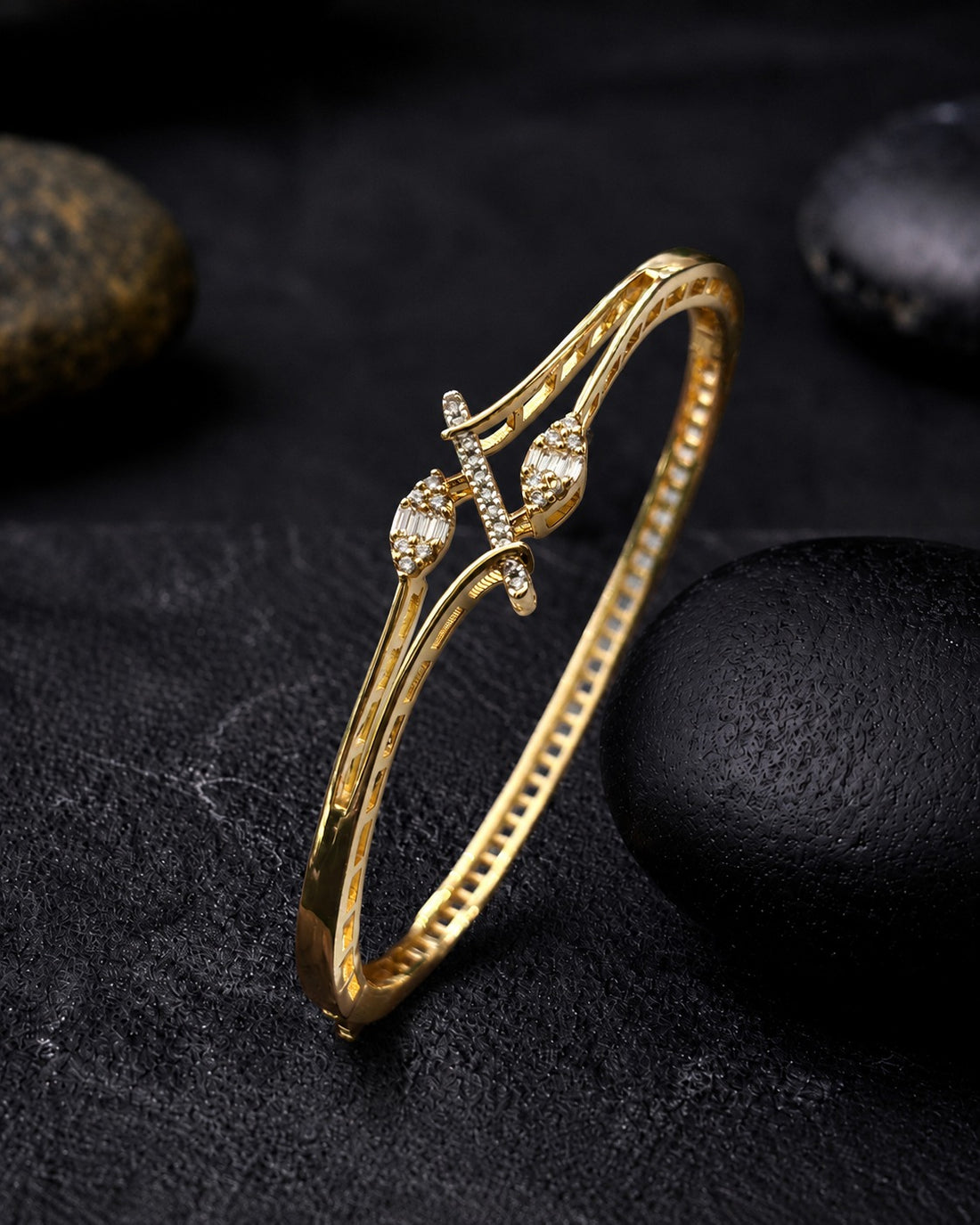 A gold-plated bangle bracelet with a decorative cross motif and small clear stones, displayed on a dark textured surface with black stones.