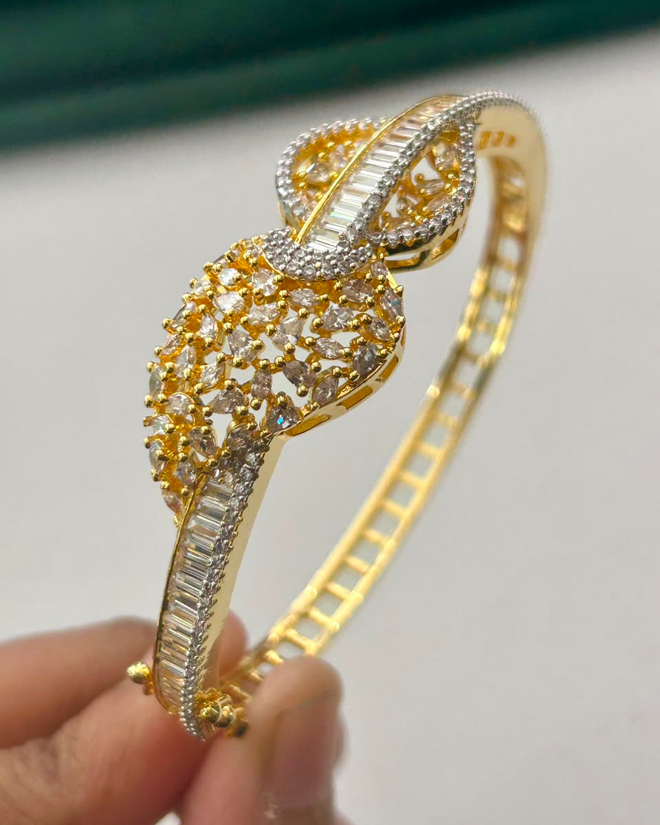 A gold-plated bangle bracelet with intricate crystal and diamond pavé detailing, held by a hand against a light gray background.