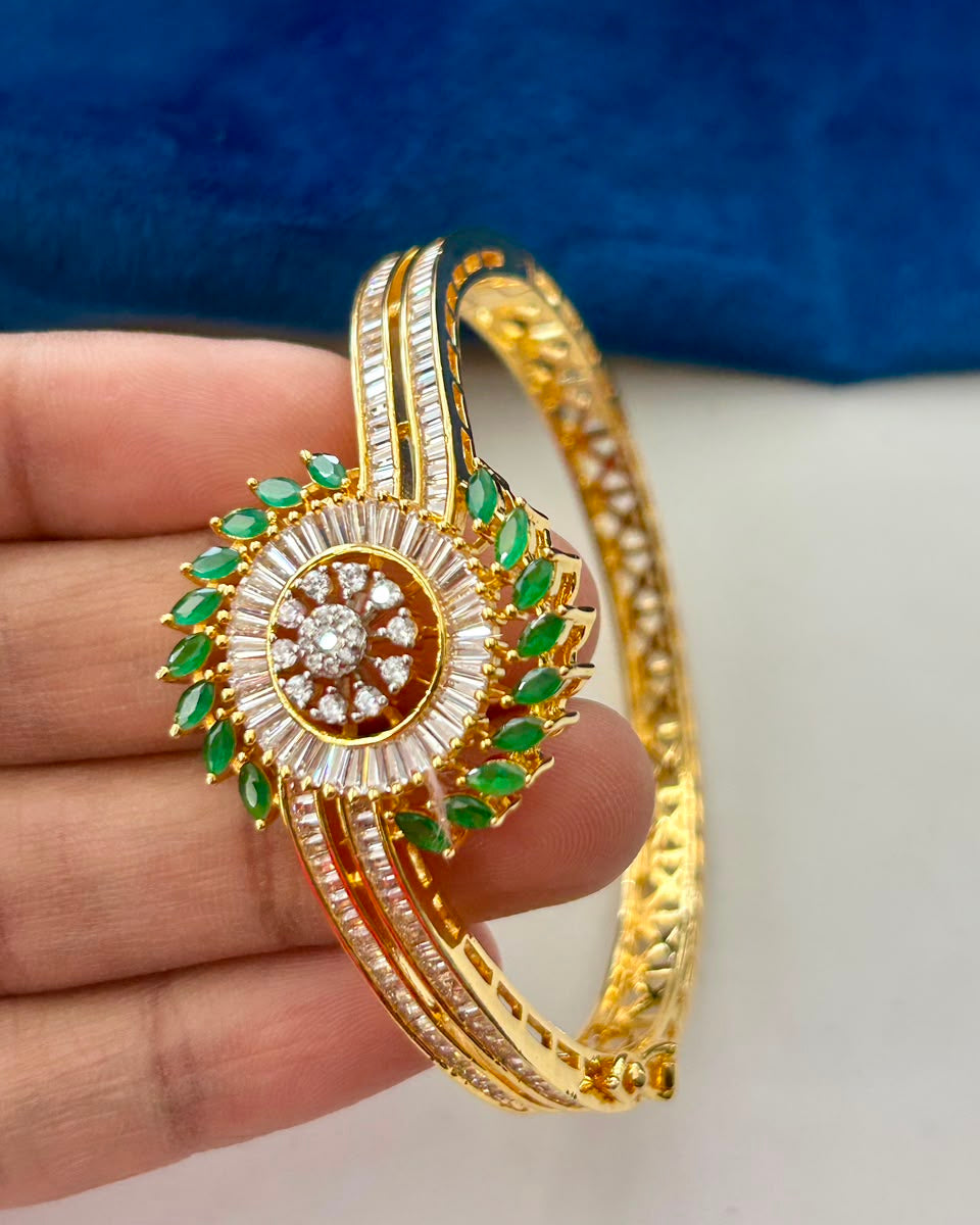 A gold-plated bangle bracelet with intricate green and white stone detailing, held in a person's hand against a blue and white background.
