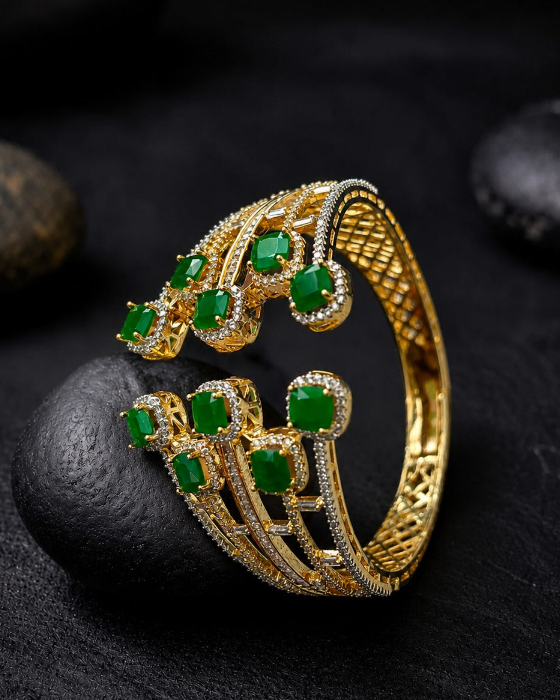 A gold-plated cuff-style ring with multiple green gemstone accents and small clear stones, displayed on a dark textured surface with smooth black stones.