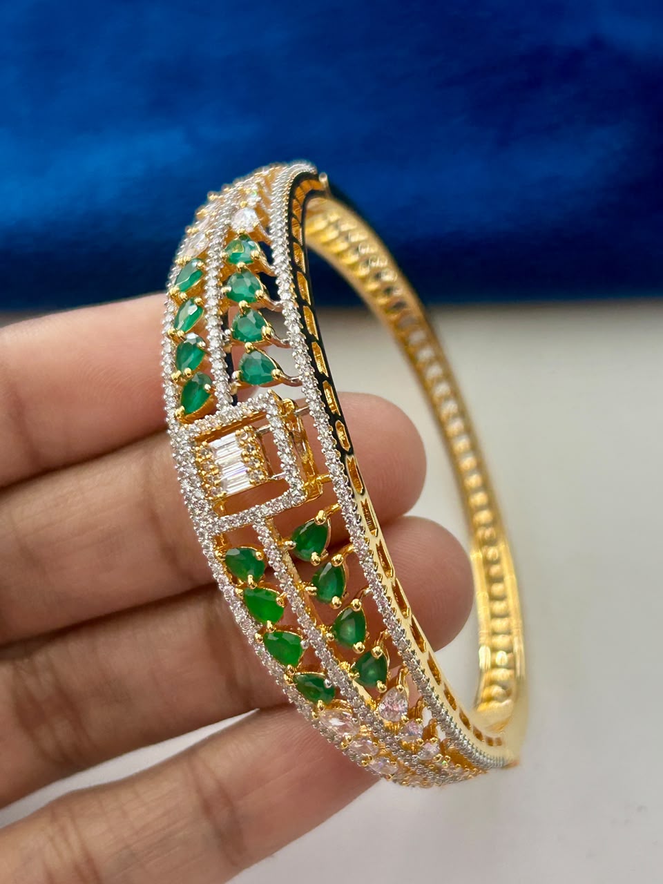 A gold-plated double-row bangle bracelet decorated with green emerald-cut gemstones and clear pavé-set stones, held in a hand against a blue velvet background.