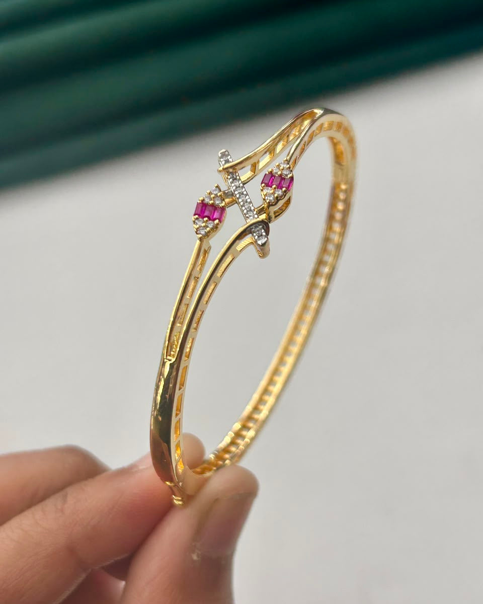A gold-plated hinged bangle bracelet with a central decorative cross motif featuring pink gemstones and small clear stones, held by a hand against a white background with a dark green blurred backdrop.
