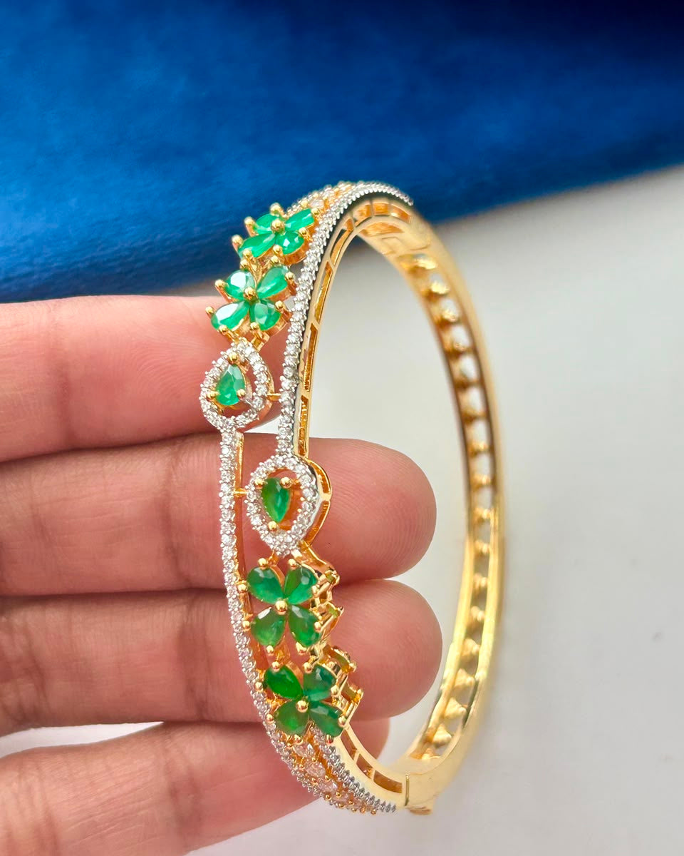 A gold-plated hinged bangle bracelet decorated with green emerald-cut stones and pavé-set clear crystals, held in a hand against a blue and white background.