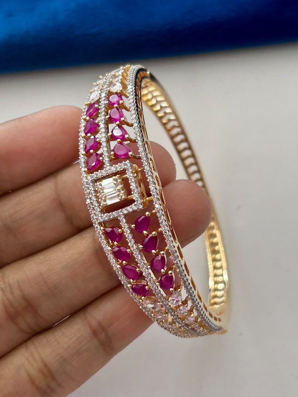 A gold-plated hinged bangle bracelet decorated with pink teardrop-cut gemstones and clear pavé-set stones, held in a person's hand against a white and blue background.