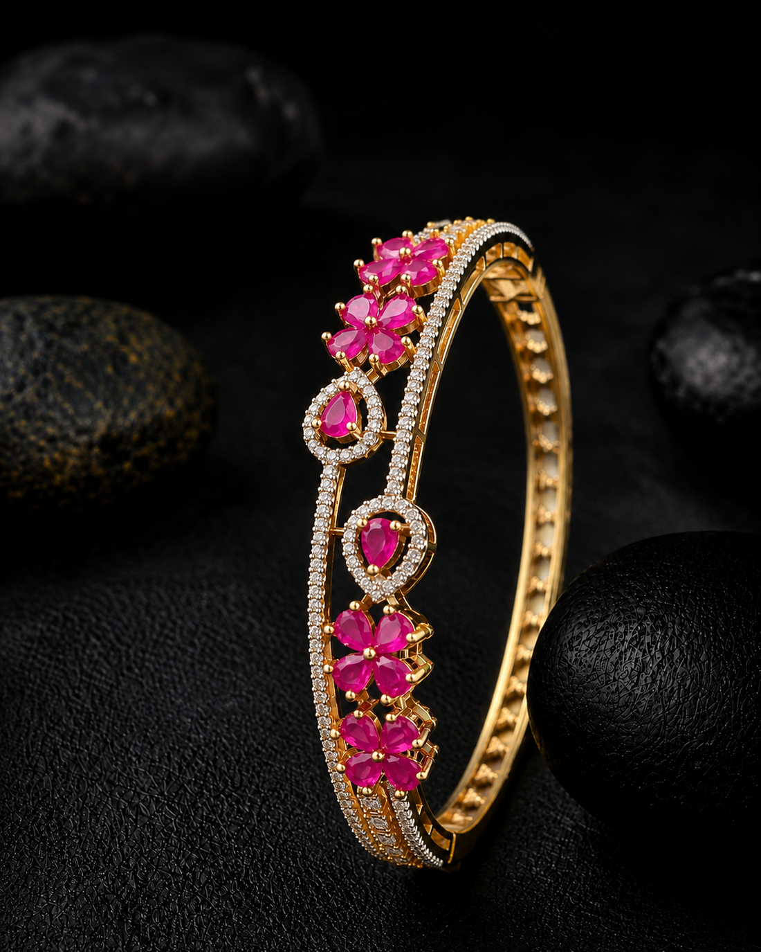 A gold-plated hinged bangle bracelet decorated with vibrant pink gemstones and clear pavé-set stones, displayed on a dark textured background with black stones.