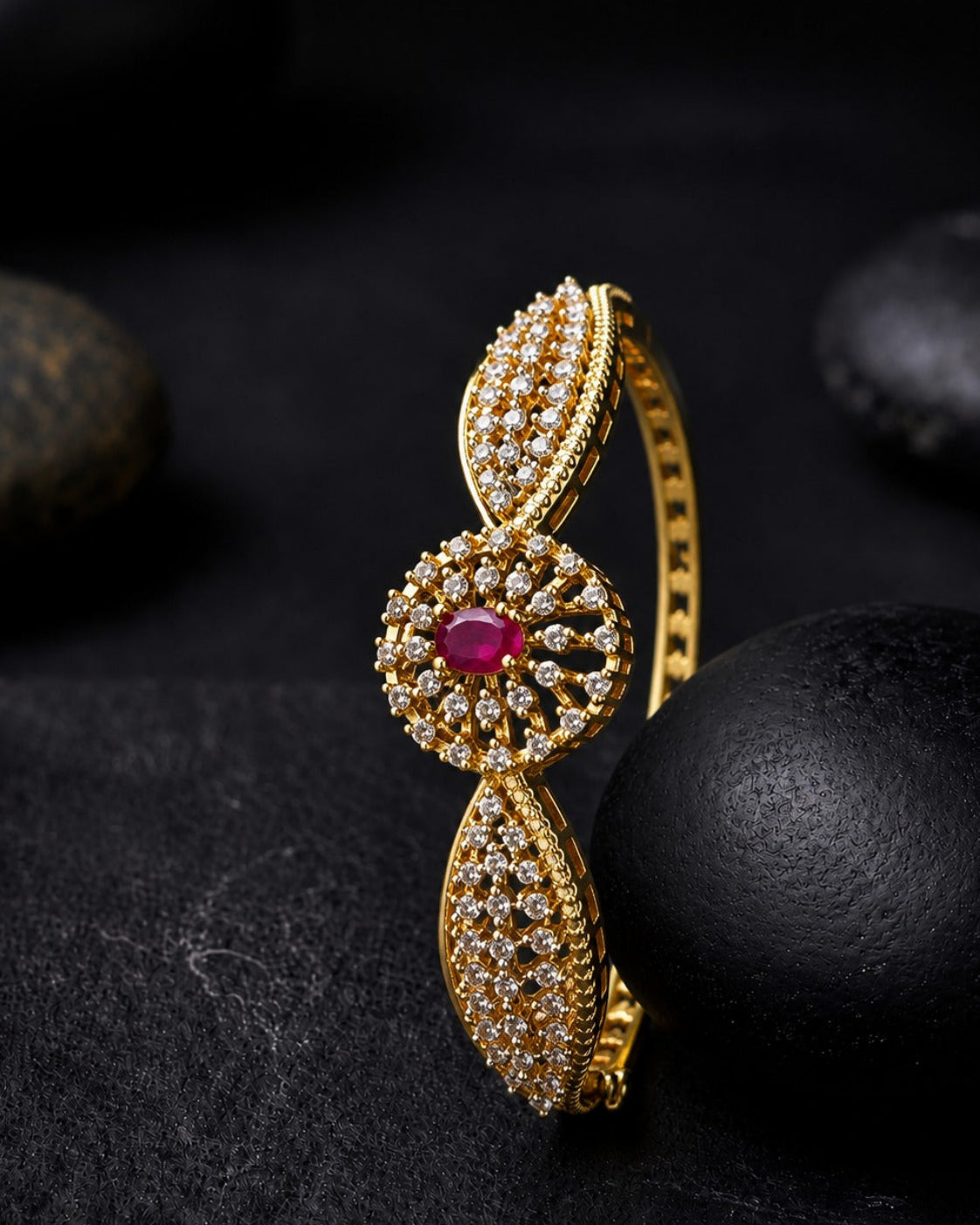 A gold-plated hinged bangle bracelet featuring a central circular motif with a vibrant pinkish-red oval ruby gemstone surrounded by small clear stones, flanked by two leaf-shaped sections paved with clear stones. The bracelet is displayed on a dark textured surface with smooth black stones in the background.