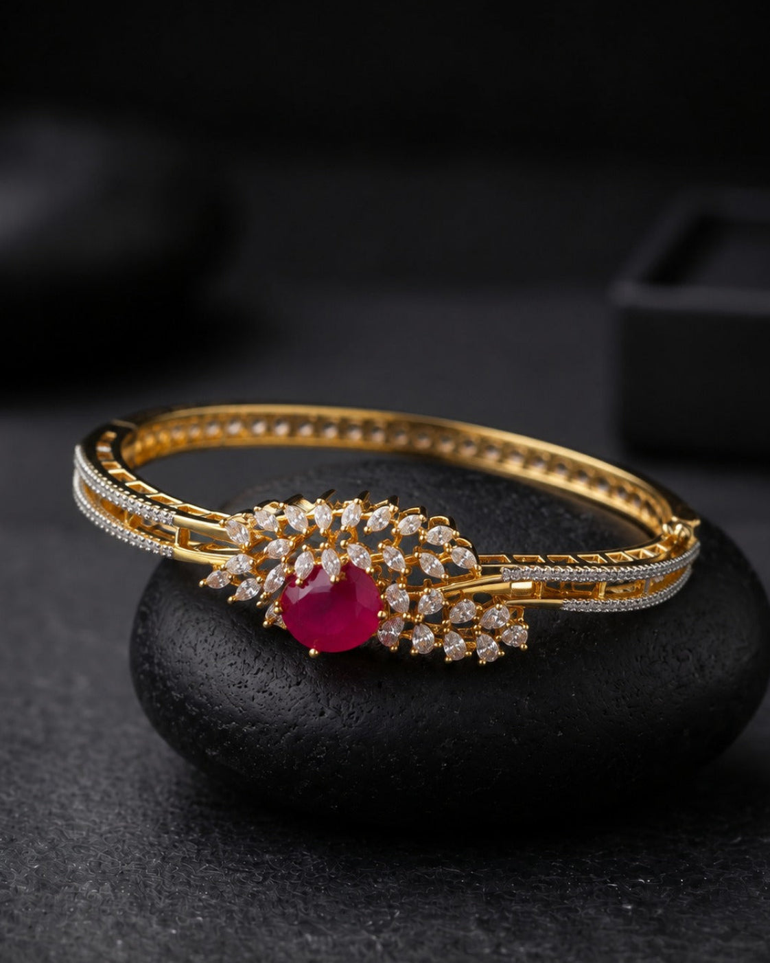 A gold-plated hinged bangle bracelet with a central floral cluster featuring a vibrant pinkish-red heart-shaped gemstone and small clear stones, resting on a dark stone prop.