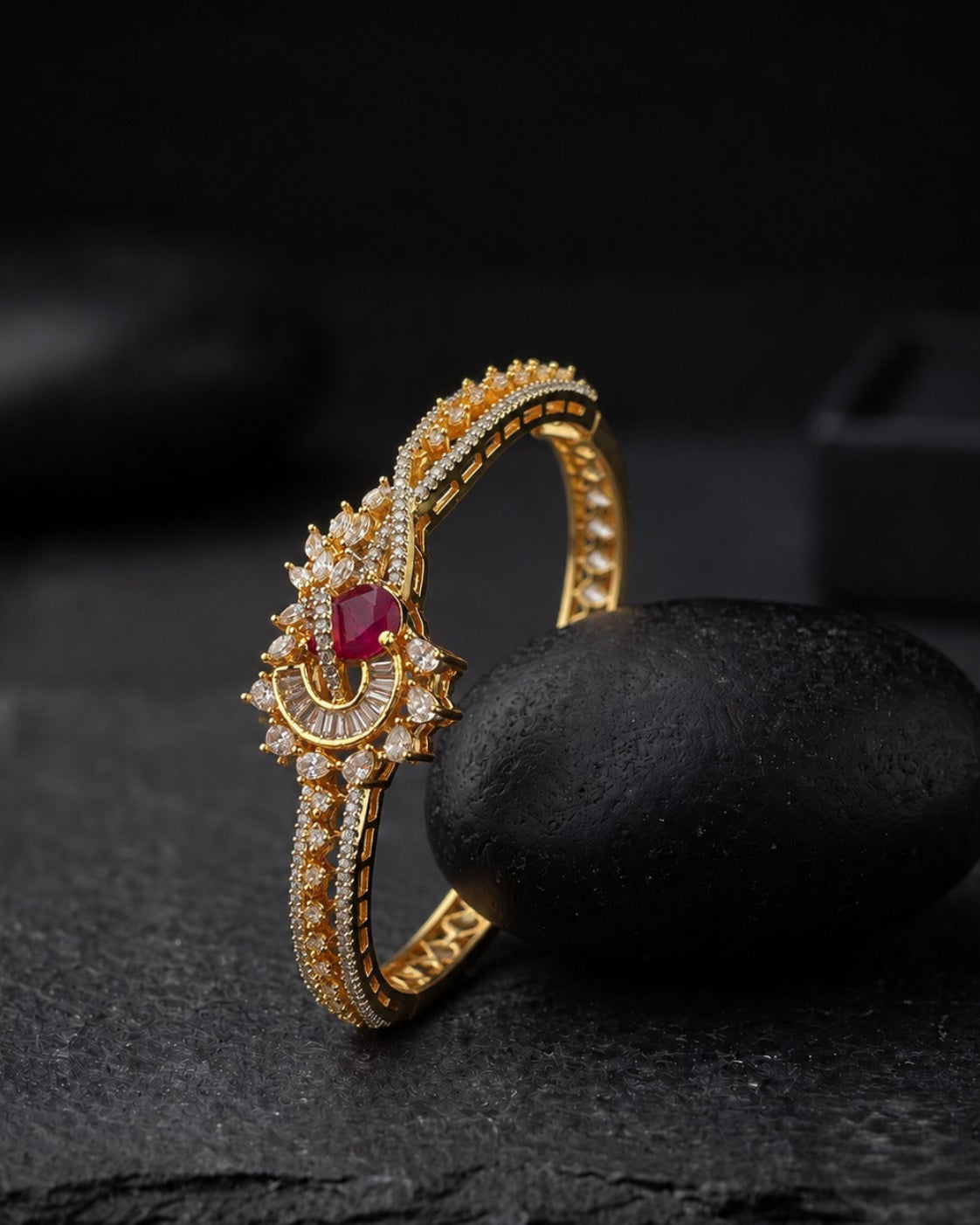 A gold-plated huggie ring with a central red oval gemstone and clear pavé accents, displayed on a dark stone prop against a dark background.