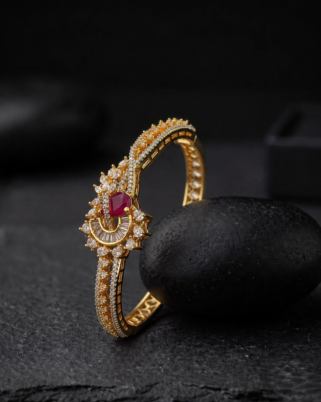 A gold-plated huggie ring with a central red oval gemstone and clear pavé accents, displayed on a dark stone prop against a dark background.