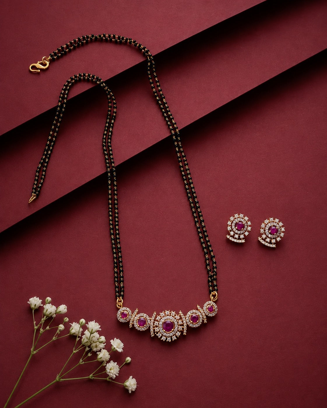 A gold-plated mangalsutra and matching stud earrings set with pinkish-red stones and surrounded by clear crystals, displayed on a dark red background with decorative baby's breath flowers.