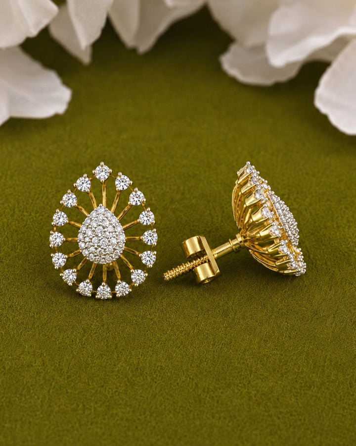 A pair of gold teardrop-shaped stud earrings adorned with clear pavé stones, shown on a textured olive green background with white flower petals at the top.