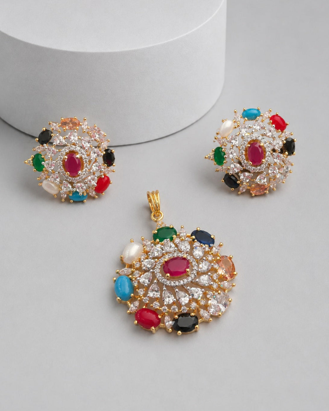 A gold-tone jewelry set featuring a large pendant and two matching stud earrings, decorated with multiple colorful gemstones including red, green, blue, black, and white pearls, arranged on a light gray surface with a white cylindrical prop.