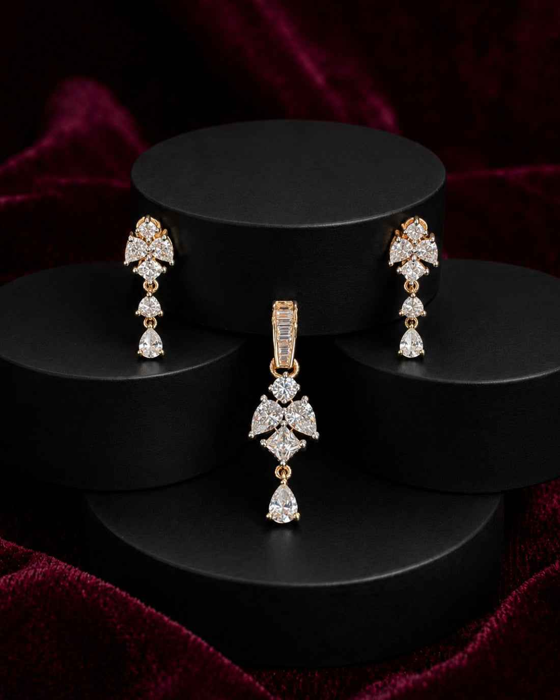 A gold-tone jewelry set featuring a matching pendant and drop earrings, both adorned with sparkling clear gemstones in a floral and teardrop design, displayed on black cylindrical pedestals against a dark red velvet background.