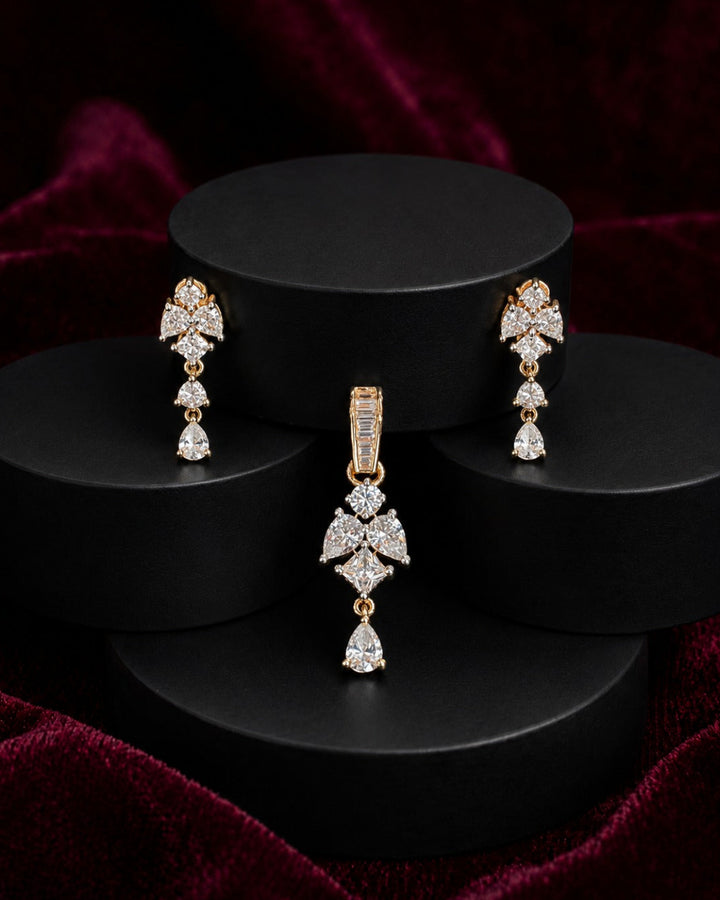 A gold-tone jewelry set featuring a matching pendant and drop earrings, both adorned with sparkling clear gemstones in a floral and teardrop design, displayed on black cylindrical pedestals against a dark red velvet background.