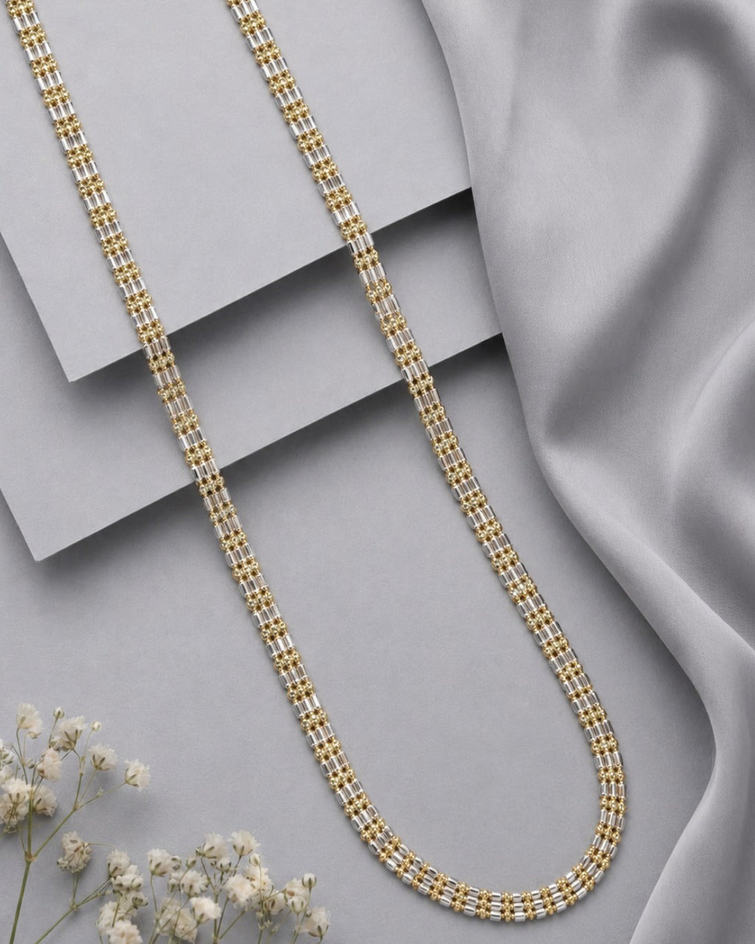 A gold-toned chain necklace with a textured, woven-link design, arranged in a U-shape on a light gray surface with layered gray cards and draped satin fabric.